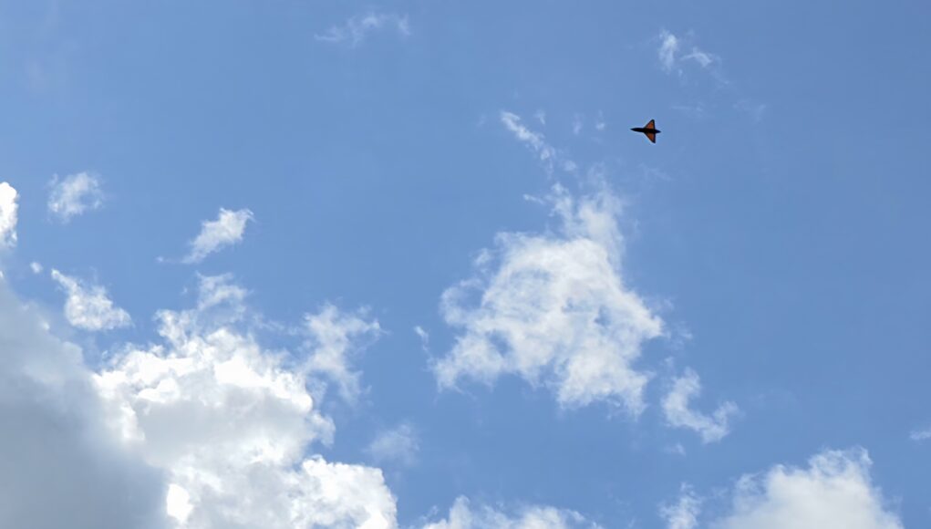 20260526_164014 Small orange delta-wing aircraft flying through a clear blue sky with a few clouds (distant plane).