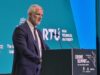 Carns warns NATO must rewire for drone war at Riga summit Man in a dark suit speaks at a podium during Drone Summit 2026; teal backdrop shows Riga Technical University and logos.
