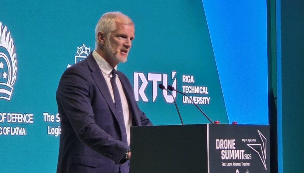 20260527_105816 Man in a dark suit speaks at a podium during Drone Summit 2026; teal backdrop shows Riga Technical University and logos.