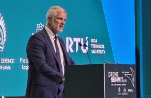 Man in a dark suit speaks at a podium during Drone Summit 2026; teal backdrop shows Riga Technical University and logos.