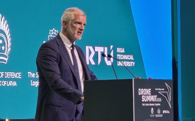 Carns warns NATO must rewire for drone war at Riga summit Man in a dark suit speaks at a podium during Drone Summit 2026; teal backdrop shows Riga Technical University and logos.