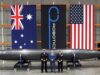 Three men in naval uniforms and a man in a blue suit stand beside a large black missile on wheeled supports in a hangar, with Australian, robotics, and US flags behind them.