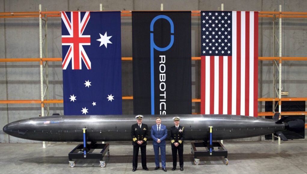 Three men in naval uniforms and a man in a blue suit stand beside a large black missile on wheeled supports in a hangar, with Australian, robotics, and US flags behind them.