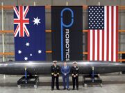 Three men in naval uniforms and a man in a blue suit stand beside a large black missile on wheeled supports in a hangar, with Australian, robotics, and US flags behind them.
