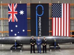 Three men in naval uniforms and a man in a blue suit stand beside a large black missile on wheeled supports in a hangar, with Australian, robotics, and US flags behind them.