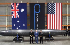 Three men in naval uniforms and a man in a blue suit stand beside a large black missile on wheeled supports in a hangar, with Australian, robotics, and US flags behind them.