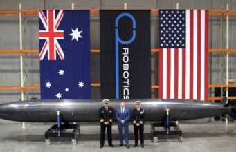 Three men in naval uniforms and a man in a blue suit stand beside a large black missile on wheeled supports in a hangar, with Australian, robotics, and US flags behind them.