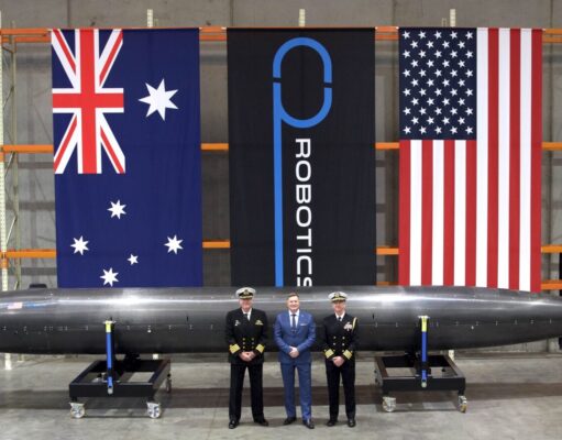 Three men in naval uniforms and a man in a blue suit stand beside a large black missile on wheeled supports in a hangar, with Australian, robotics, and US flags behind them.