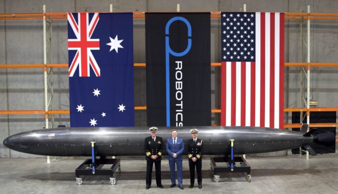 Three men in naval uniforms and a man in a blue suit stand beside a large black missile on wheeled supports in a hangar, with Australian, robotics, and US flags behind them.