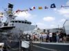 Crowd of visitors on the deck of a large gray naval ship, with colorful signal flags fluttering overhead.