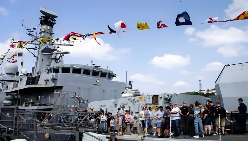 Crowd of visitors on the deck of a large gray naval ship, with colorful signal flags fluttering overhead.