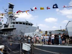 Crowd of visitors on the deck of a large gray naval ship, with colorful signal flags fluttering overhead.
