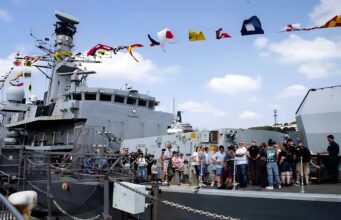 Crowd of visitors on the deck of a large gray naval ship, with colorful signal flags fluttering overhead.
