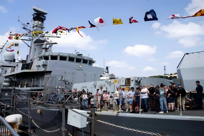 Crowd of visitors on the deck of a large gray naval ship, with colorful signal flags fluttering overhead.