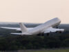 Large white commercial airliner lifting off from a runway, wheels extended, over a forested landscape as it climbs into the sky.