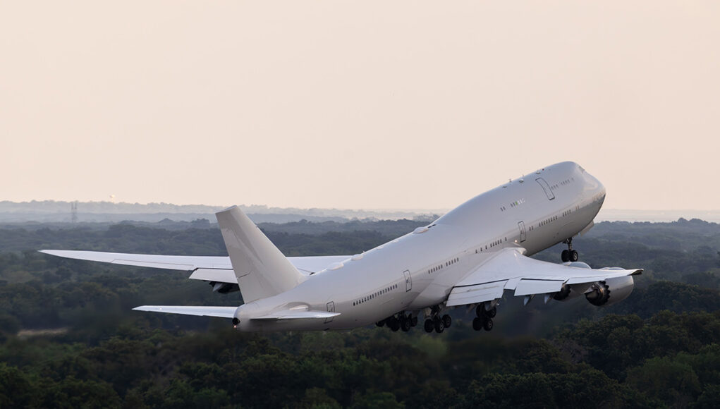 Large white commercial airliner lifting off from a runway, wheels extended, over a forested landscape as it climbs into the sky.