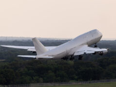 Large white commercial airliner lifting off from a runway, wheels extended, over a forested landscape as it climbs into the sky.