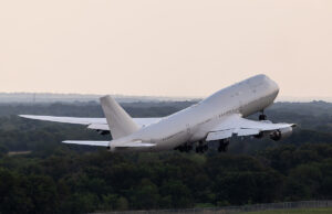 Large white commercial airliner lifting off from a runway, wheels extended, over a forested landscape as it climbs into the sky.