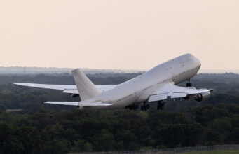 Large white commercial airliner lifting off from a runway, wheels extended, over a forested landscape as it climbs into the sky.