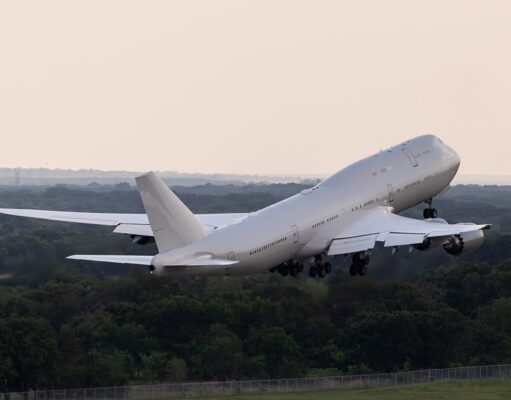 Large white commercial airliner lifting off from a runway, wheels extended, over a forested landscape as it climbs into the sky.