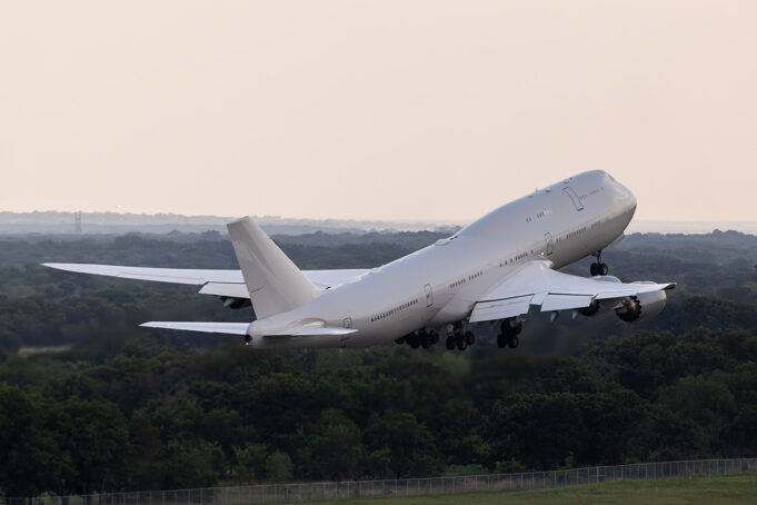 Large white commercial airliner lifting off from a runway, wheels extended, over a forested landscape as it climbs into the sky.