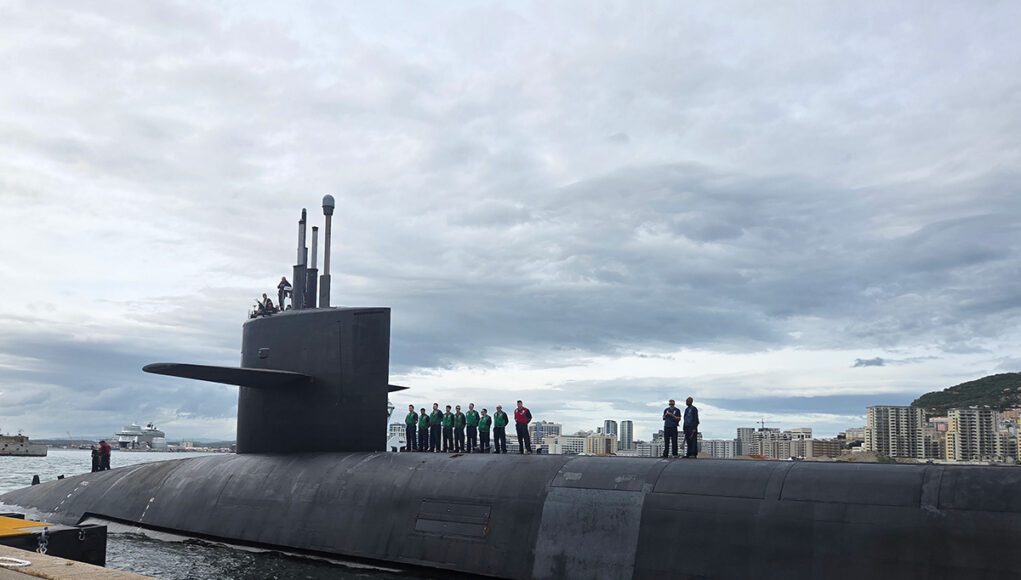 Submarine moored at the quay with crew on its deck and a city skyline in the background.
