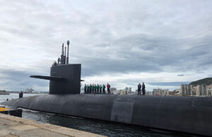Submarine moored at the quay with crew on its deck and a city skyline in the background.