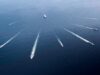 Aerial view of several motorboats leaving white wake trails across deep blue ocean surface (six boats visible).