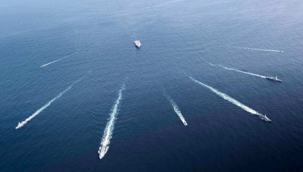 Aerial view of several motorboats leaving white wake trails across deep blue ocean surface (six boats visible).