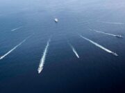 Aerial view of several motorboats leaving white wake trails across deep blue ocean surface (six boats visible).