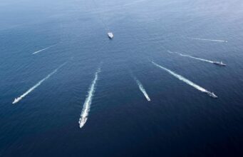 Aerial view of several motorboats leaving white wake trails across deep blue ocean surface (six boats visible).