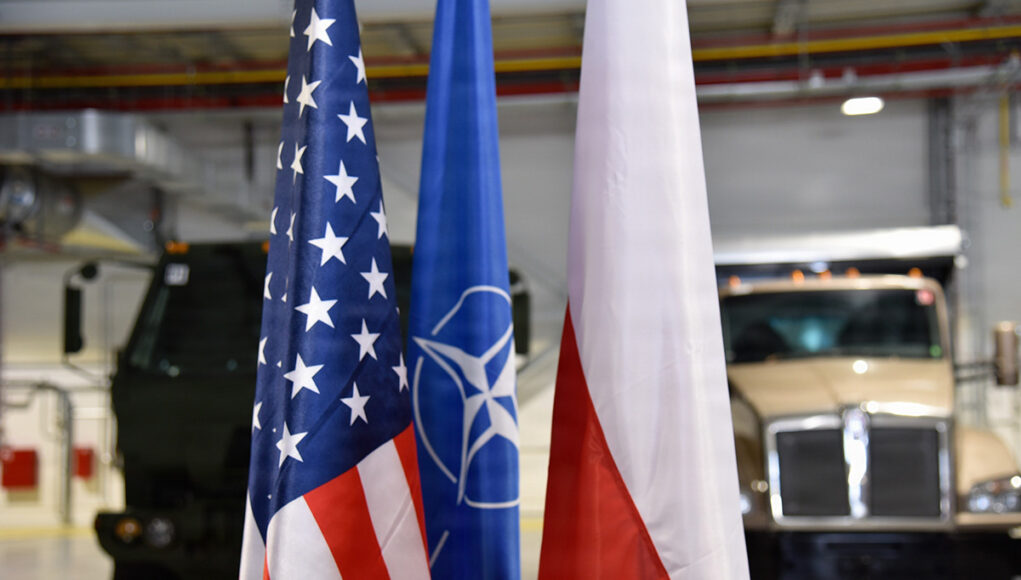 260527-A-WZ074-1001 Flags of the United States, NATO, and Poland in front of military trucks inside a warehouse or hangar.