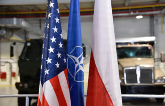 US and Poland complete deployable air base storage site Flags of the United States, NATO, and Poland in front of military trucks inside a warehouse or hangar.