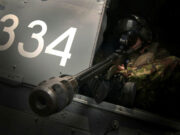 Belgian firm to acquire British rifle maker Military helicopter side with large white '334' numbers and a crewman in camouflage sitting at a mounted gun in the open hatch, aiming outward.