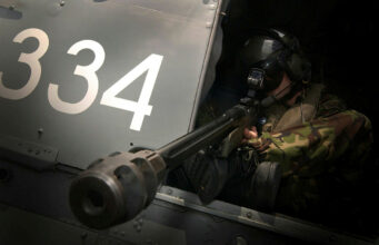 Military helicopter side with large white '334' numbers and a crewman in camouflage sitting at a mounted gun in the open hatch, aiming outward.