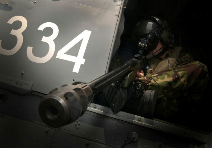 Military helicopter side with large white '334' numbers and a crewman in camouflage sitting at a mounted gun in the open hatch, aiming outward.