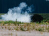 Smoke rising from a dark, rusted barbecue in a grassy field, with a tree line in the distance.