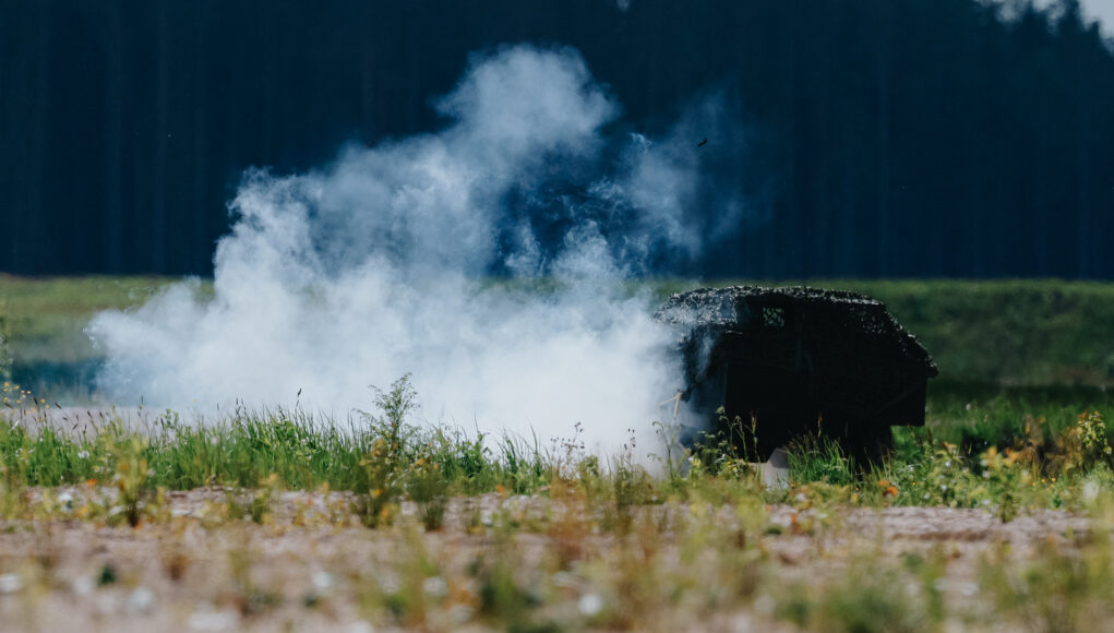 Smoke rising from a dark, rusted barbecue in a grassy field, with a tree line in the distance.