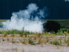 Smoke rising from a dark, rusted barbecue in a grassy field, with a tree line in the distance.