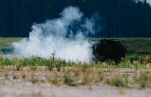 Smoke rising from a dark, rusted barbecue in a grassy field, with a tree line in the distance.