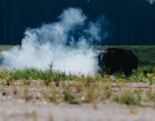 Smoke rising from a dark, rusted barbecue in a grassy field, with a tree line in the distance.