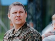 Man in camouflage military uniform looking left outdoors, with another person partially visible.
