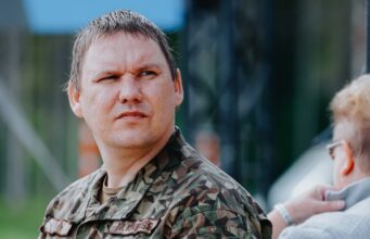Man in camouflage military uniform looking left outdoors, with another person partially visible.