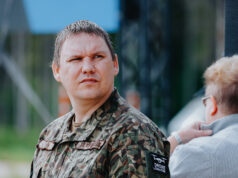 Man in camouflage military jacket with a serious expression, outdoors near a fence; someone else touches his shoulder in the background.