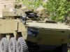 Tan armored fighting vehicle with turret-mounted cannon and many wheels parked beside a stone wall and green trees