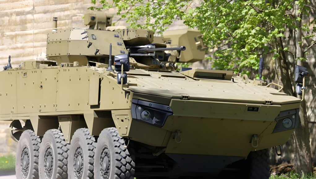 Tan armored fighting vehicle with turret-mounted cannon and many wheels parked beside a stone wall and green trees