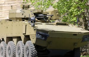 Tan armored fighting vehicle with turret-mounted cannon and many wheels parked beside a stone wall and green trees