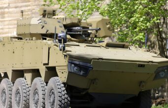 Tan armored fighting vehicle with turret-mounted cannon and many wheels parked beside a stone wall and green trees