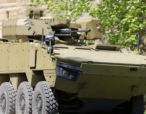 Tan armored fighting vehicle with turret-mounted cannon and many wheels parked beside a stone wall and green trees
