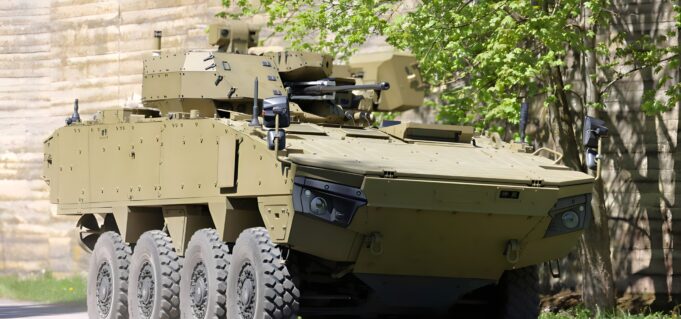 Tan armored fighting vehicle with turret-mounted cannon and many wheels parked beside a stone wall and green trees